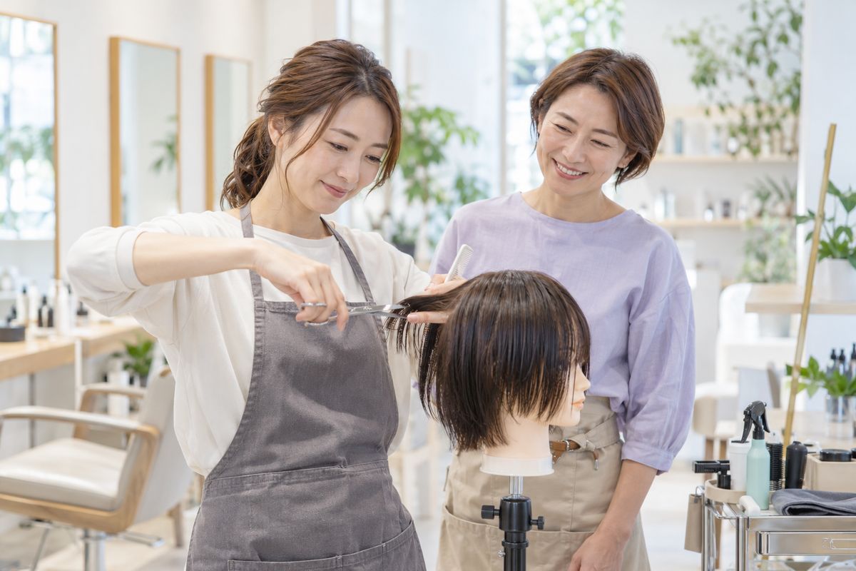 復帰する美容師がウィッグで練習し、先輩が見守るイメージ