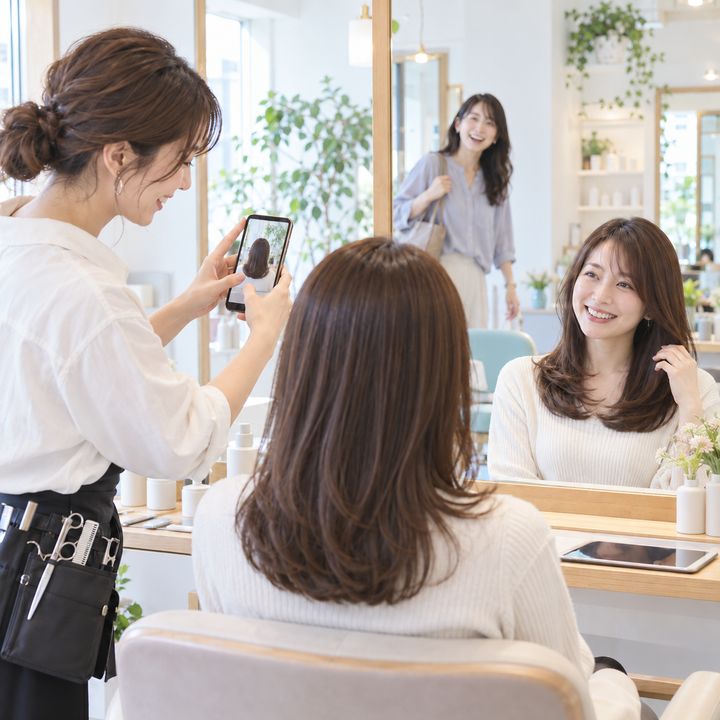 仕上がった髪をスマートフォンで撮影し、お客様と笑顔を交わす美容師のイメージ